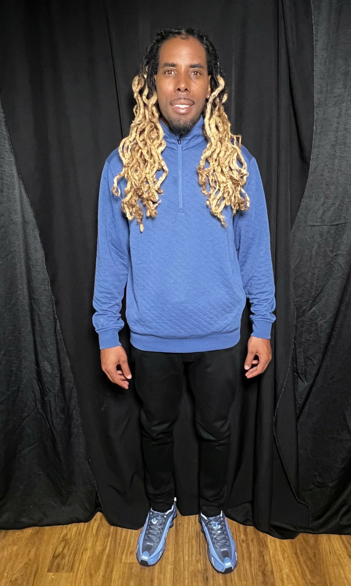 Person with long curly hair wearing a blue sweatshirt, black pants, and blue sneakers, standing in front of black curtains.