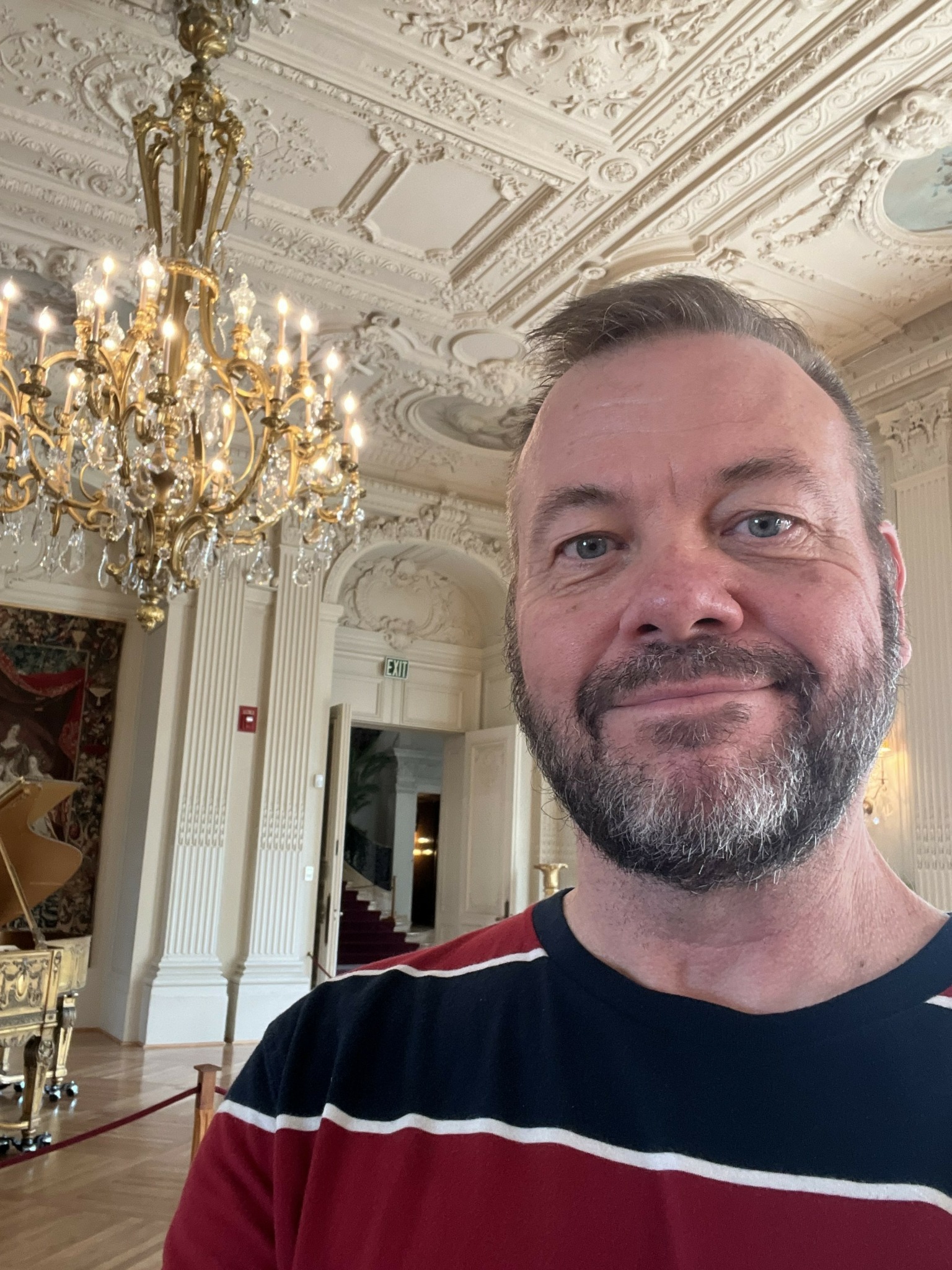 Man with beard smiling in ornate room with chandelier and detailed ceiling.