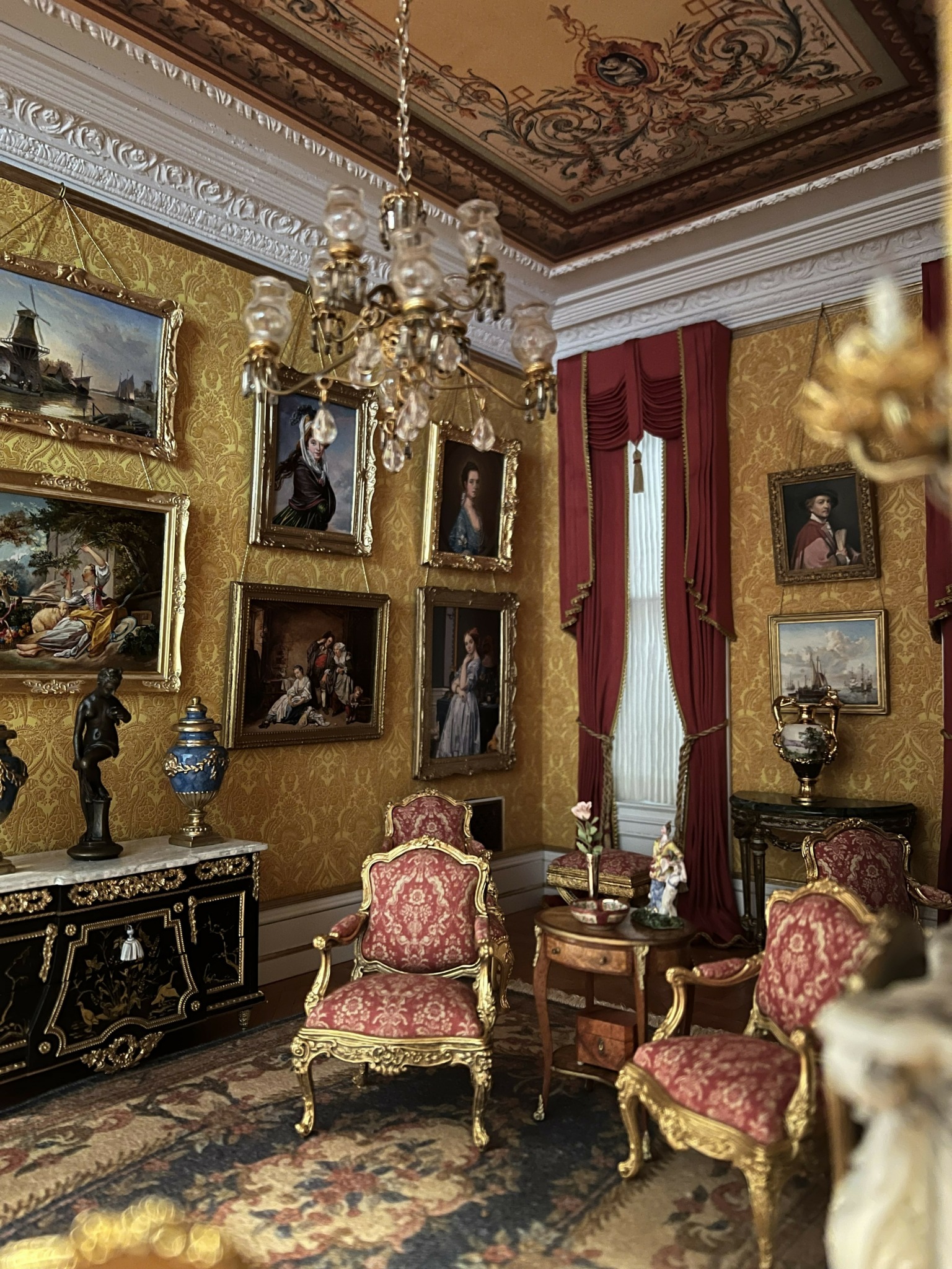 Elegant room with gold framed portraits, ornate furniture, chandelier, and red curtains, decorated in a classical style.