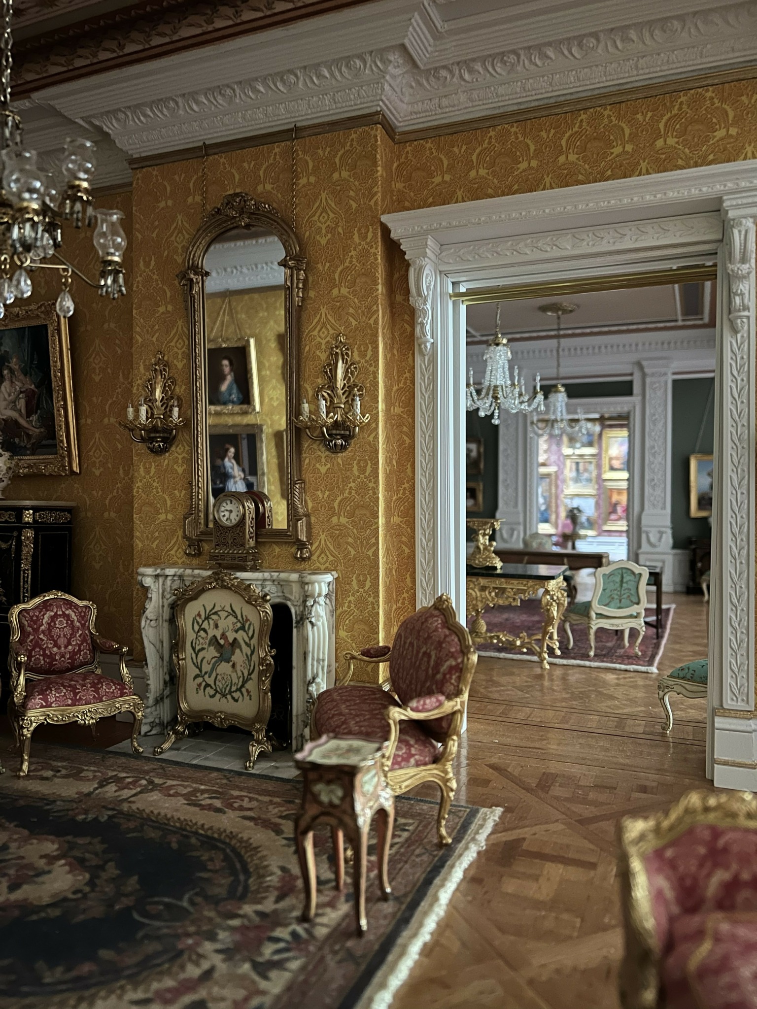 Elegant room with antique furniture, ornate mirror, chandelier, and patterned carpet, leading to a bright, decorated sitting area.