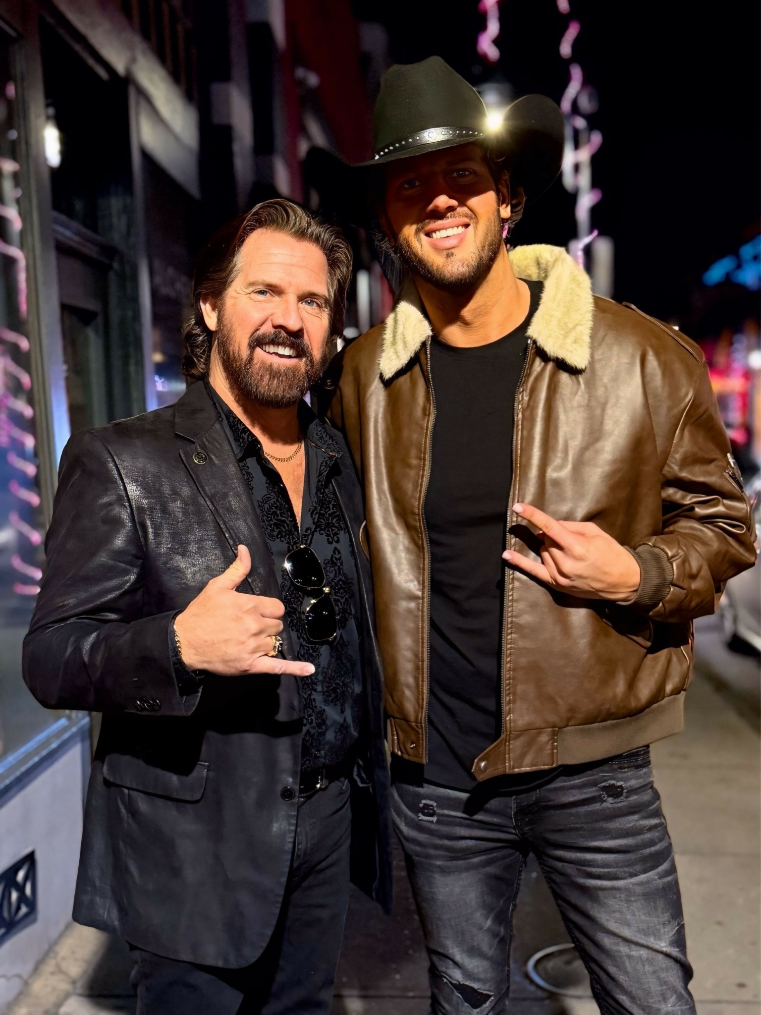 Two men standing outdoors at night, smiling, one making a hand gesture, wearing jackets and sunglasses, city lights in background.