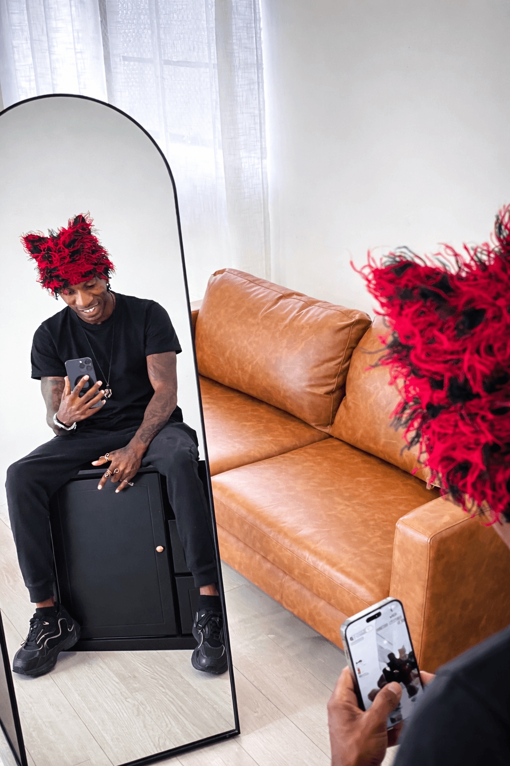 Person wearing a red feathered headpiece taking a selfie in a mirror, sitting on a black suitcase, with a brown sofa nearby.
