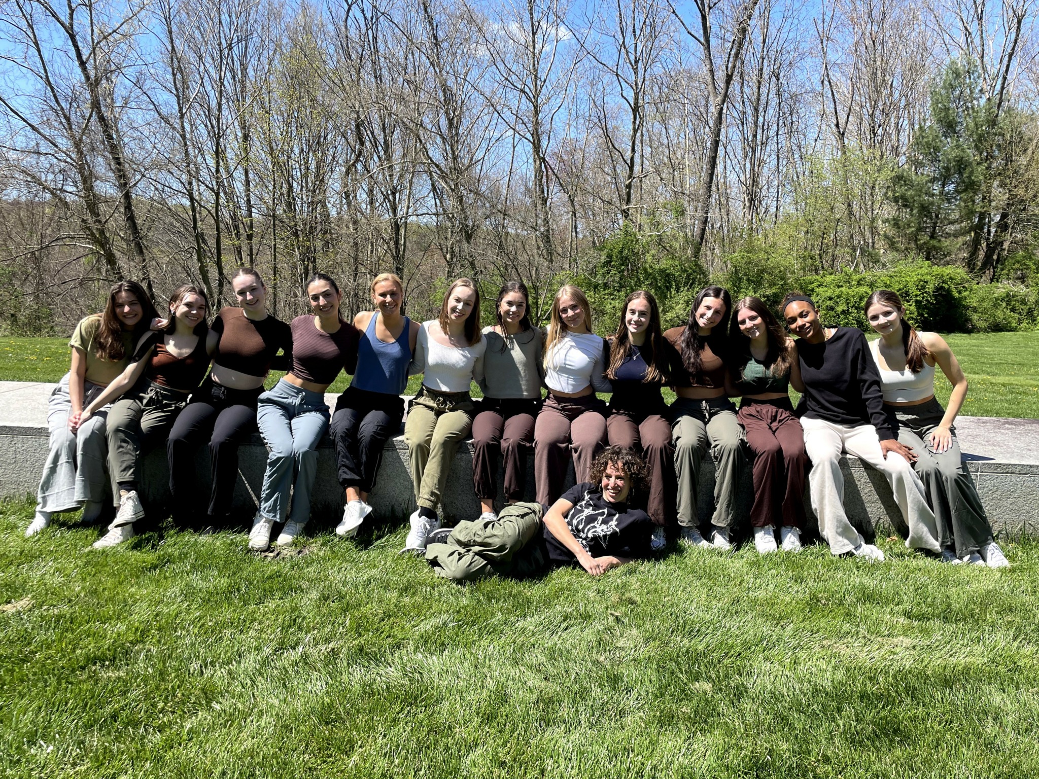 Group of people sitting on grass outdoors with trees and blue sky in background.