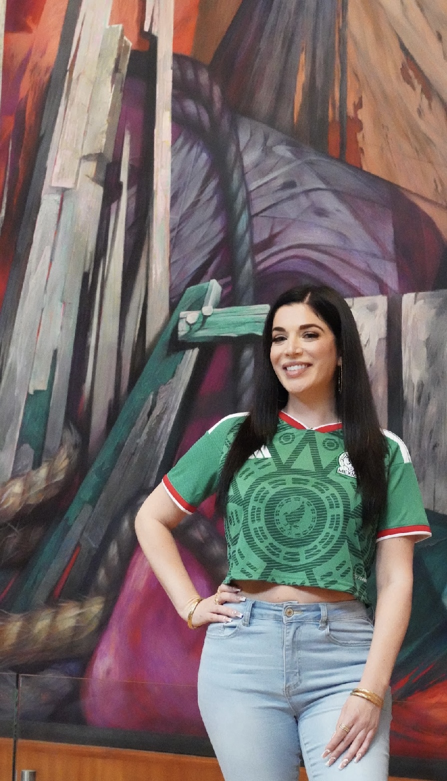 Young woman with long dark hair smiling, wearing a green patterned top and light blue jeans, standing in front of a colorful mural.
