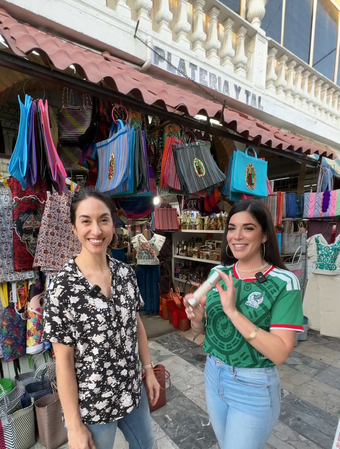 Two women smiling in front of a market stall with colorful bags and accessories, one wearing a black and white floral shirt, the other in a green shirt and jeans.