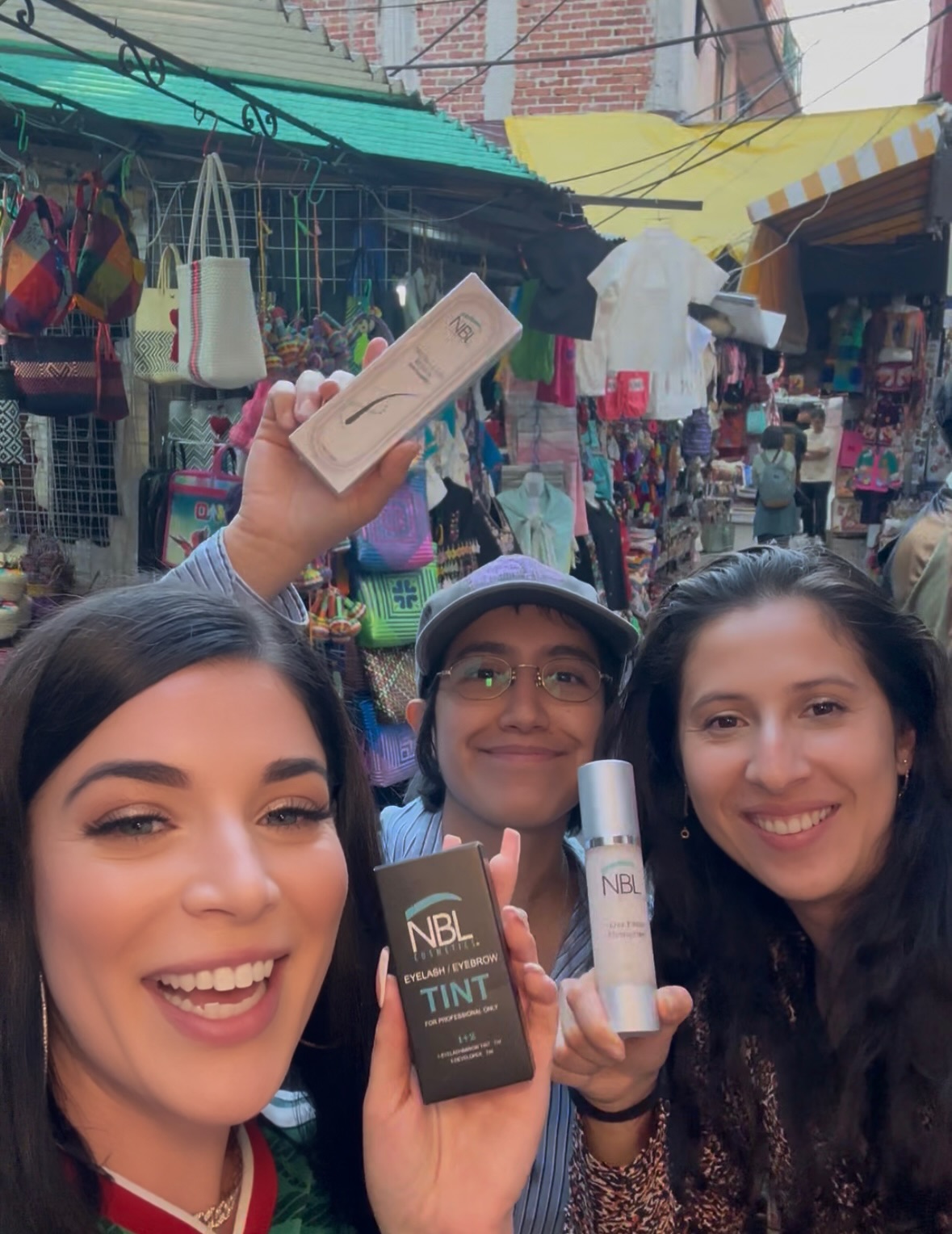 Three women smiling at a market, holding beauty products, with colorful stalls and hanging bags in the background.