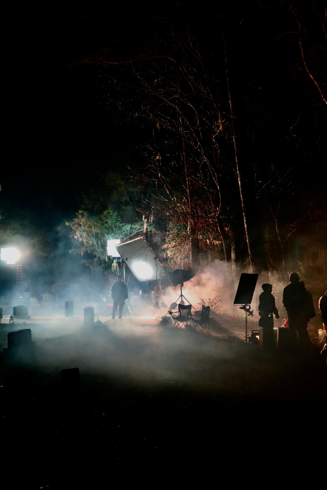 People gathered outdoors at night with smoke and fireworks, some standing and others sitting, illuminated by bright lights.