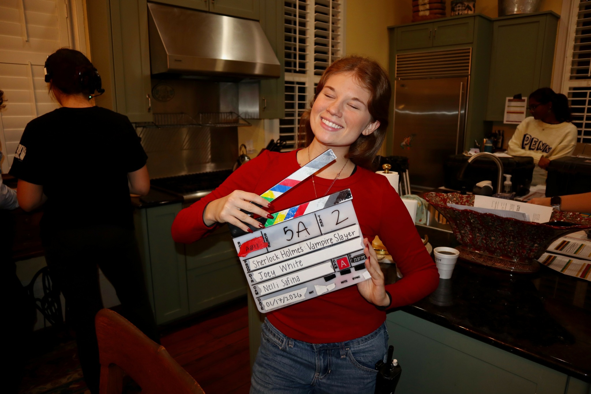 Woman smiling and holding a clapperboard in a kitchen setting, with other people in the background.