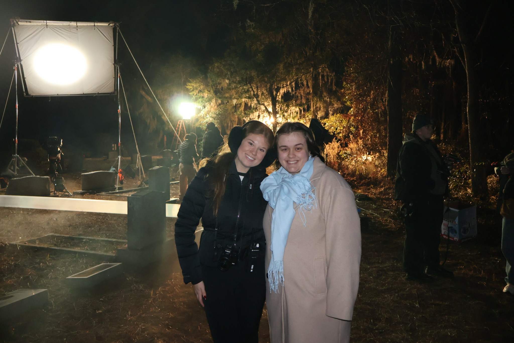 Two women standing outdoors at night, smiling, with a lit background and equipment, surrounded by trees and other people.