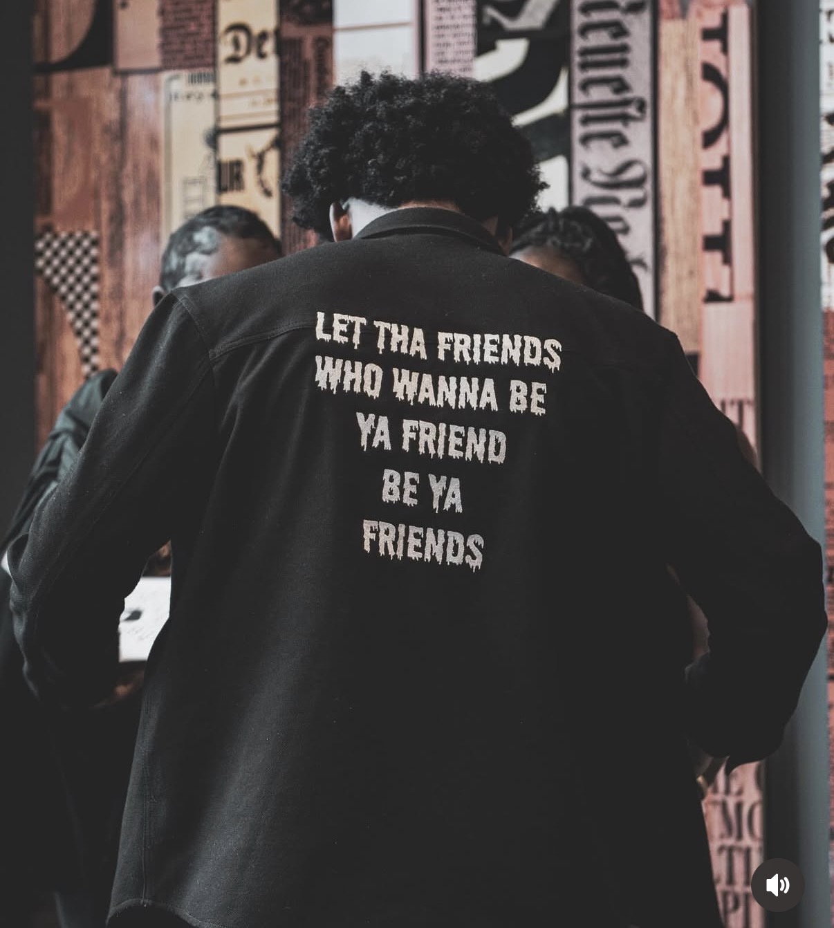Person wearing a black jacket with white text on the back, in front of a colorful background with text and patterns.