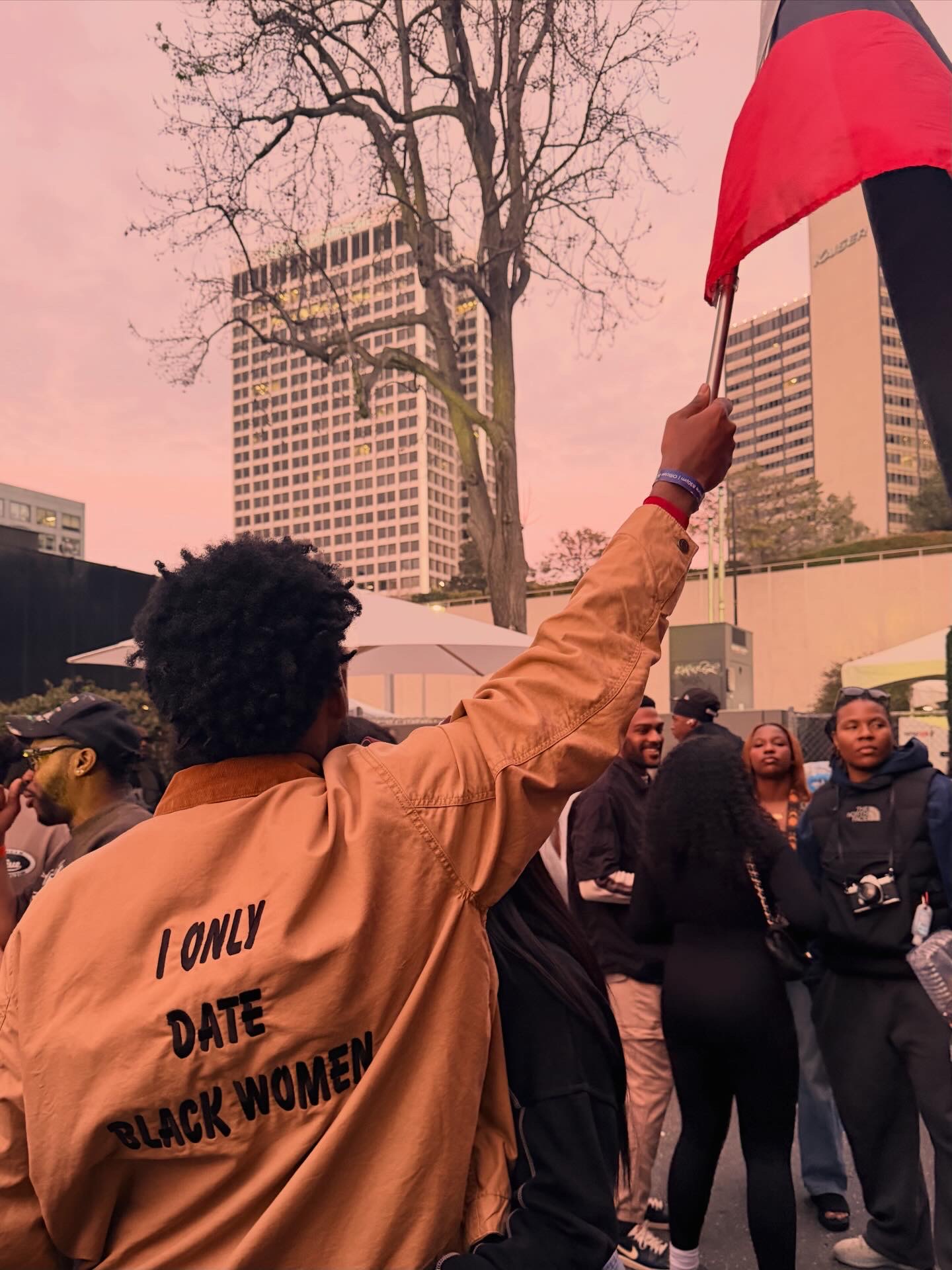 Person holding a red flag in a crowd outdoors with tall buildings and a leafless tree in the background.