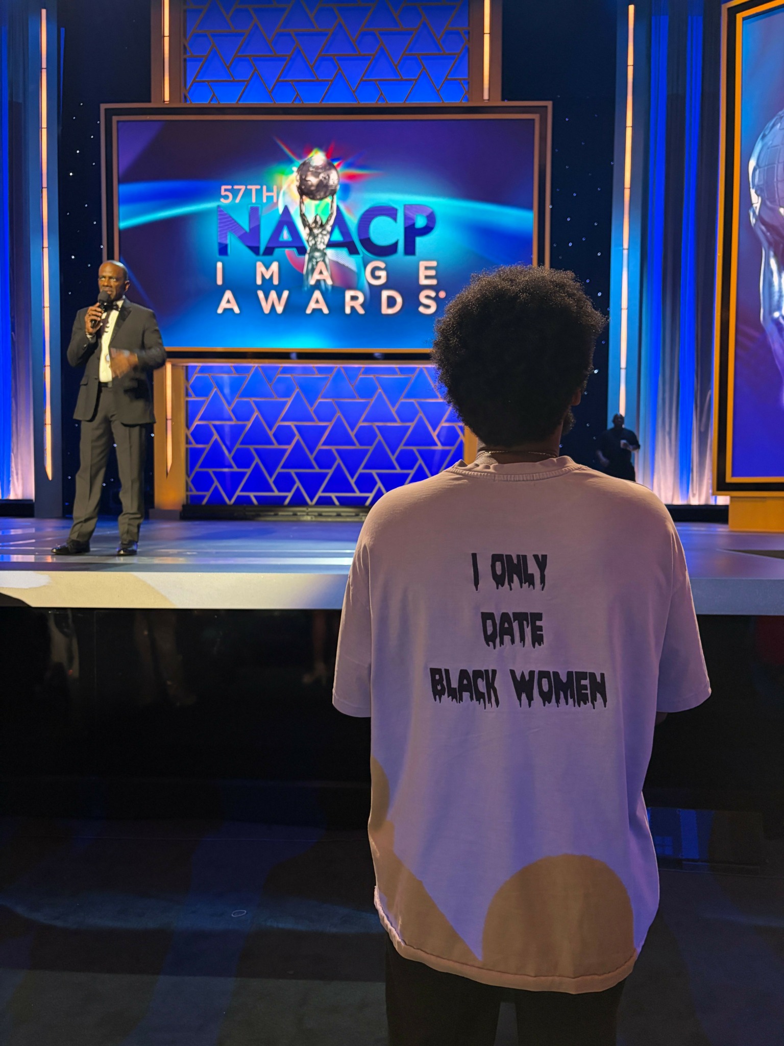 Person with curly hair wearing a white T-shirt with text, standing in front of a stage with a large screen displaying 'NAACP IMAGE AWARDS' and a host on stage.