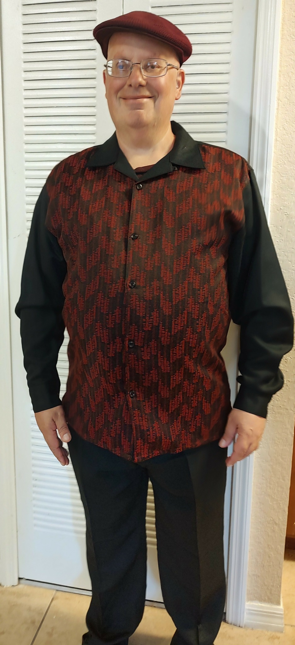 Person wearing a red hat, glasses, black and red patterned shirt, standing indoors in front of a white door.