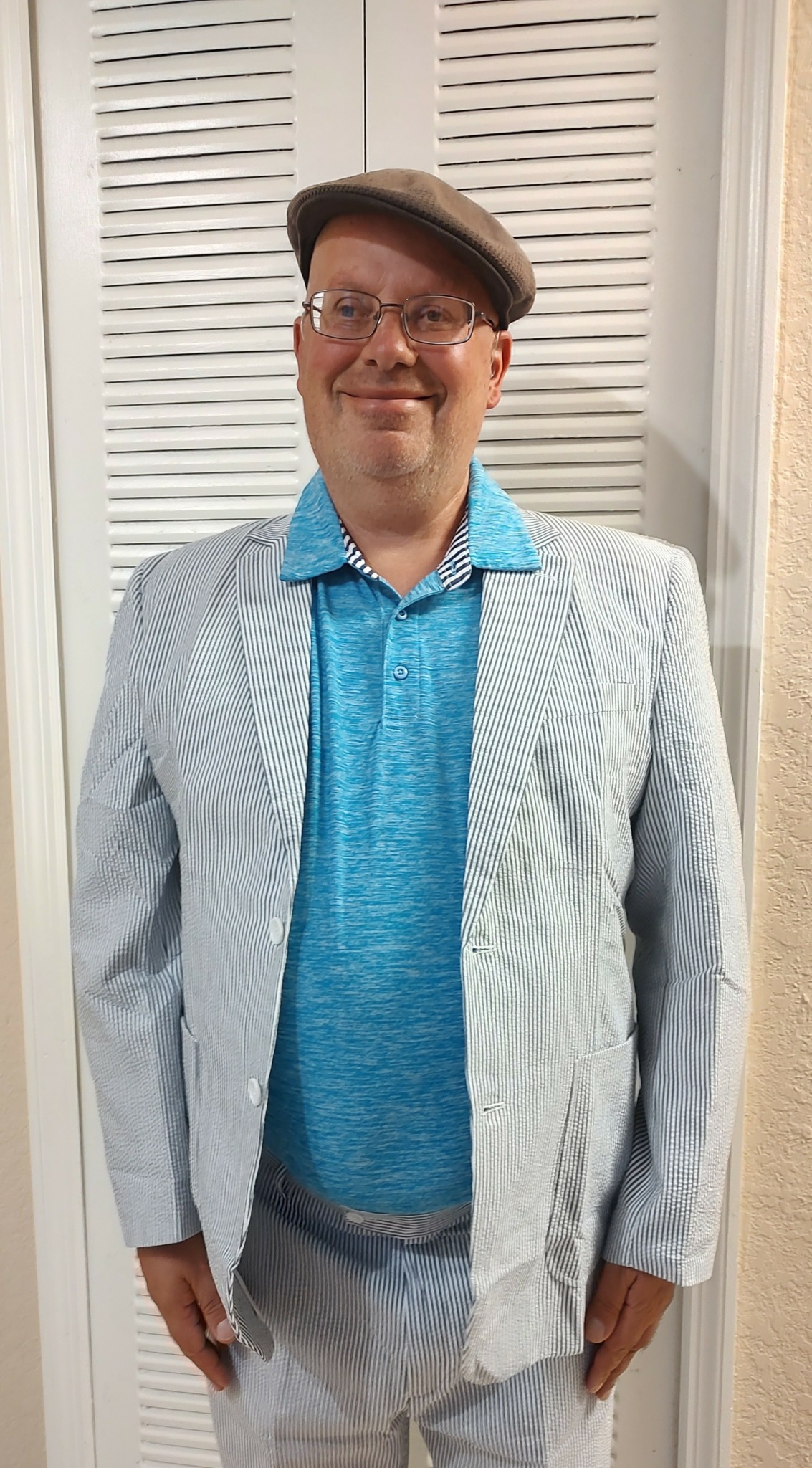 Man wearing glasses, a flat cap, a blue shirt, and a light striped blazer, standing in front of white louvered doors.