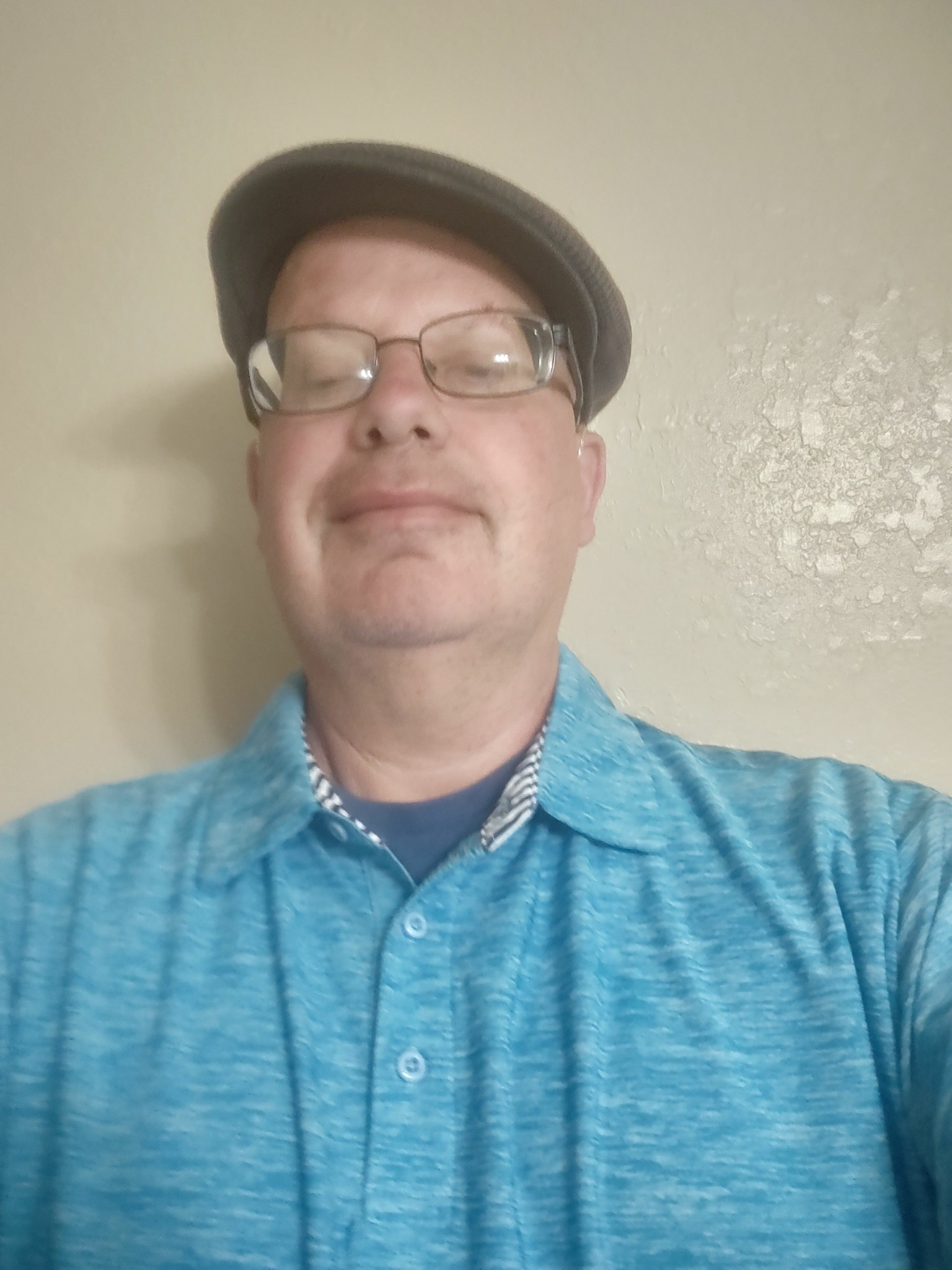 Person wearing glasses, a hat, and a blue shirt, standing indoors against a plain wall.