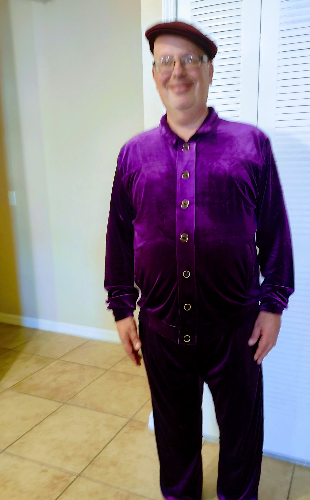 Person wearing a purple velvet shirt, glasses, and a red cap, standing indoors near a white door and beige tiled floor.