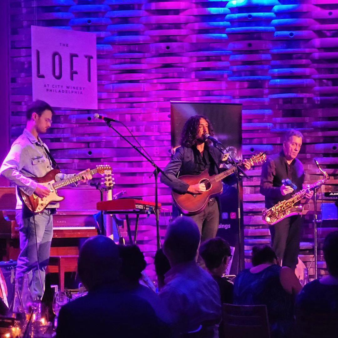 Three musicians perform on stage with guitars and saxophone, audience seated in front, purple and blue lighting, brick wall background.