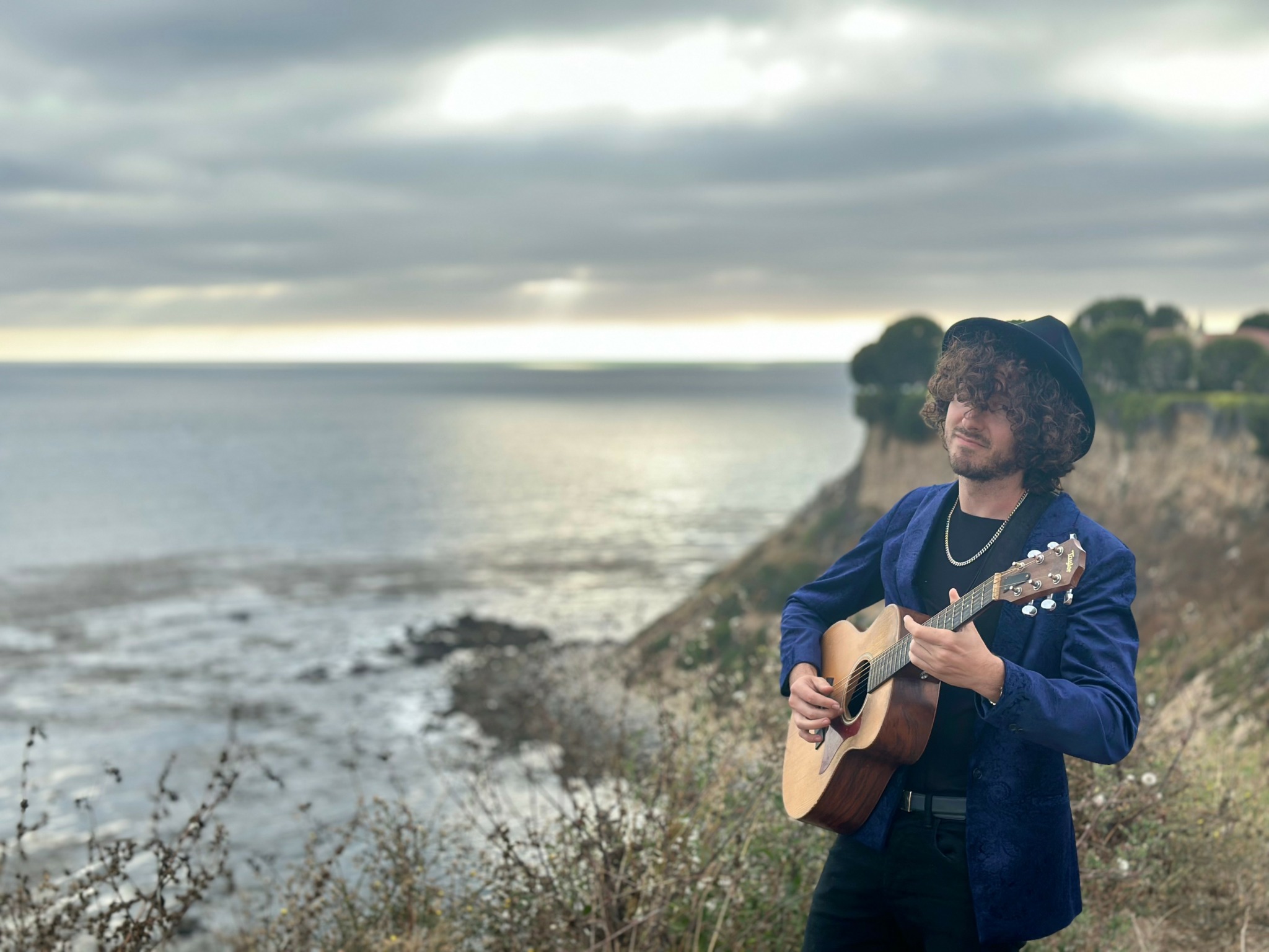 Person with curly hair playing guitar outdoors near water, cloudy sky, and grassy landscape in background.