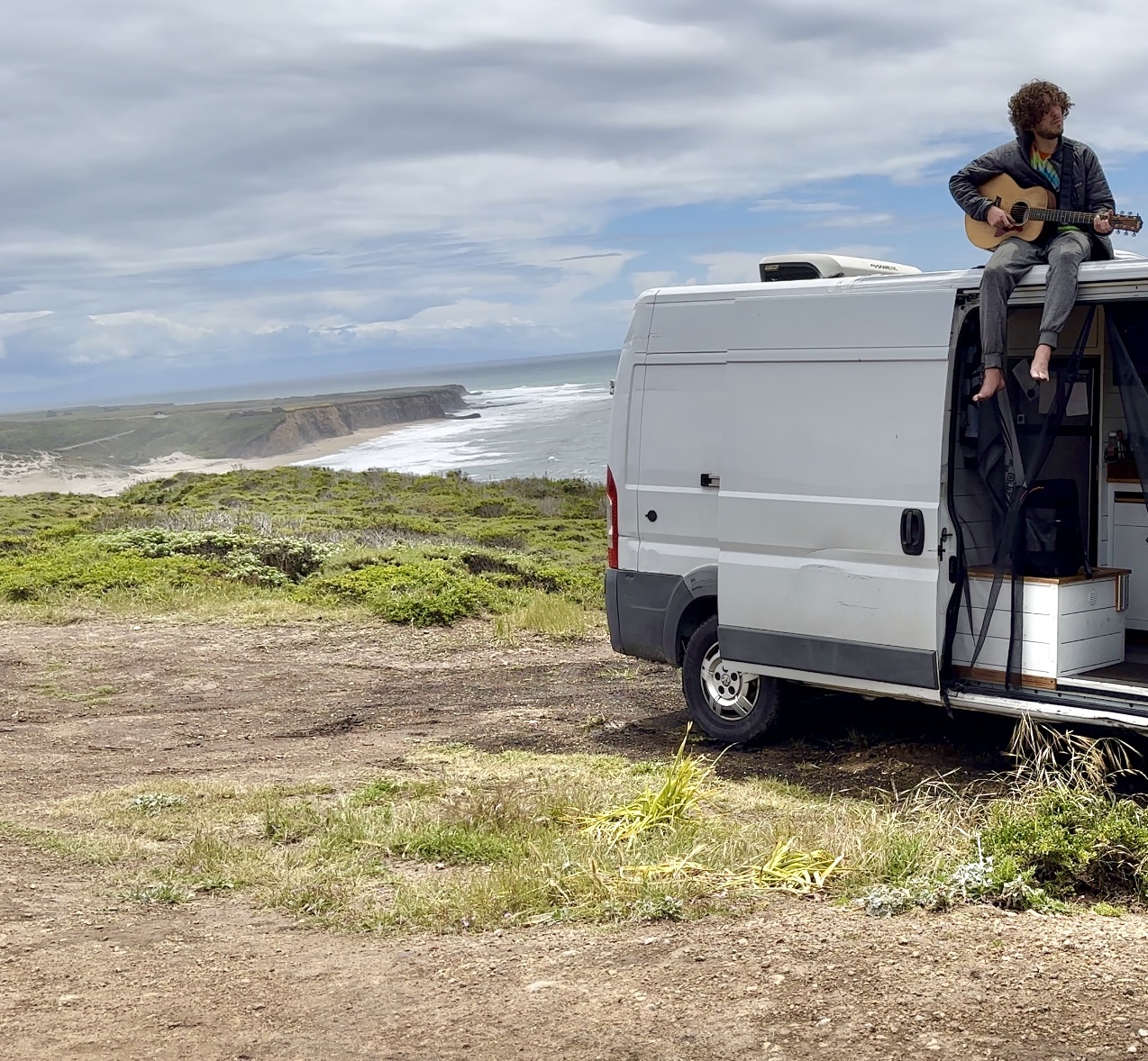 Person playing guitar on top of a white van parked near a grassy area with a coastal landscape in the background.