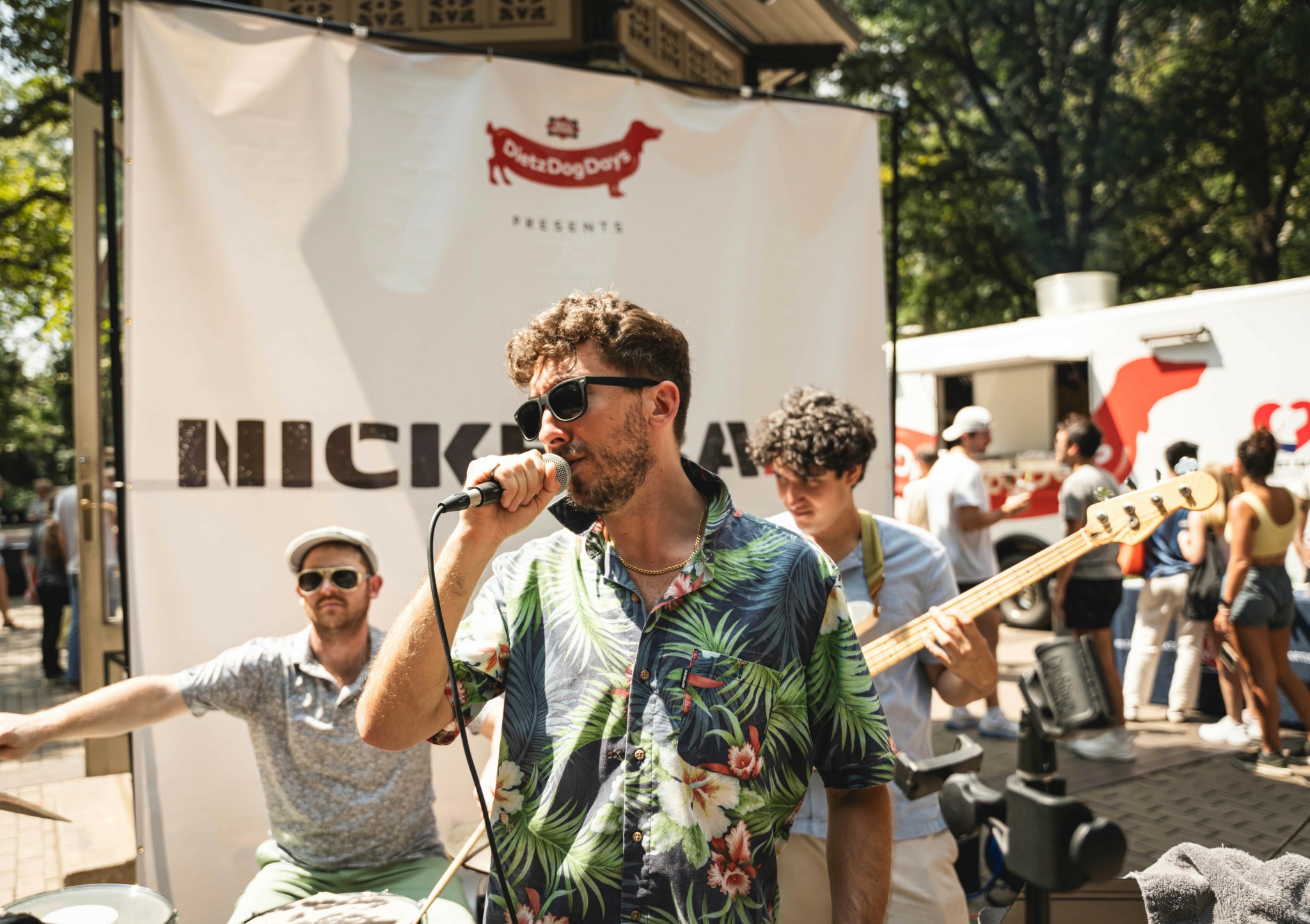 Man with sunglasses singing into a microphone outdoors, surrounded by people and trees, with a Nick logo in the background.