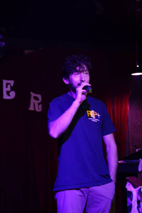 Young man with curly hair holding a microphone on stage, wearing a blue T-shirt, in a dimly lit setting.