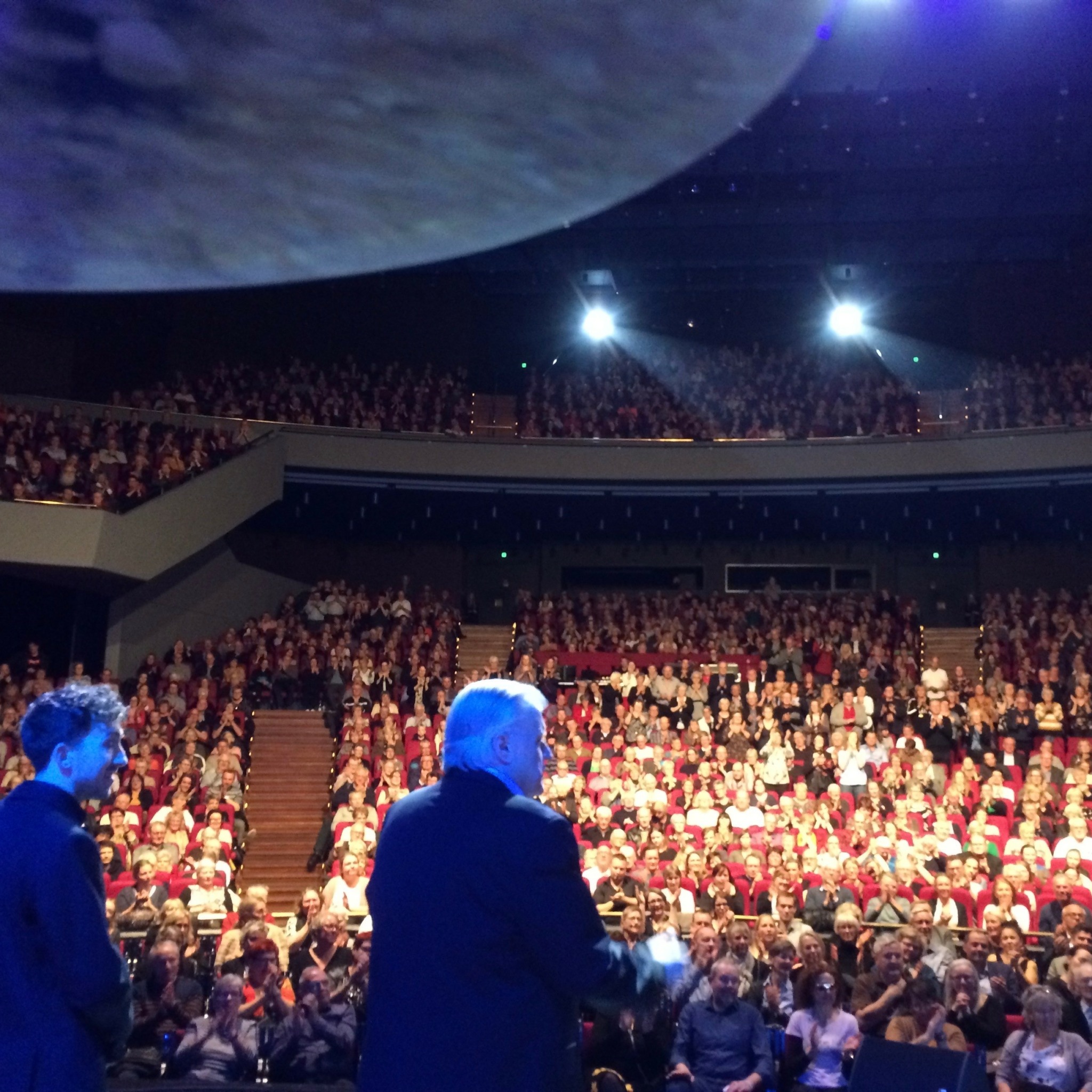 A speaker addresses a large audience in an auditorium with a view of Earth from space above.
