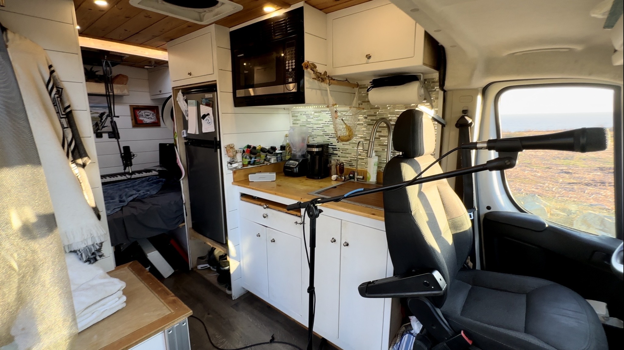 Interior of a camper van with kitchen area, microwave, and a bed visible through an open door, with a microphone setup near the driver's seat.