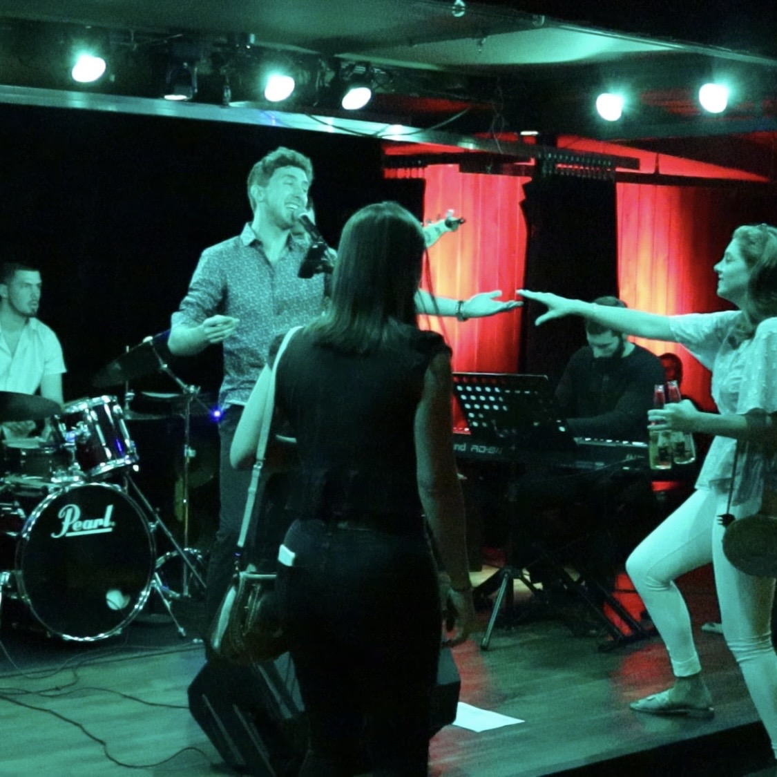 People performing on stage with musical instruments and a red curtain backdrop, under stage lighting.
