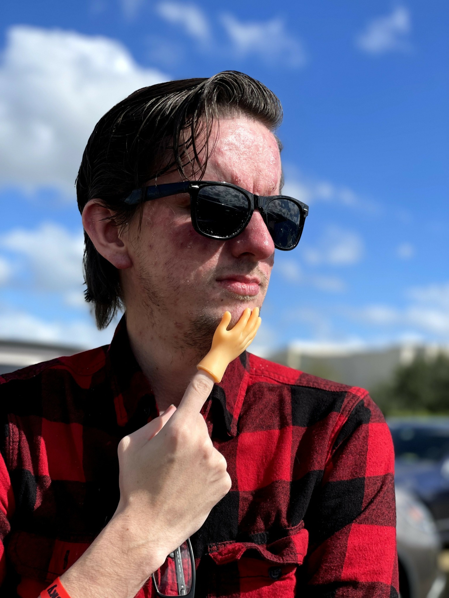 Young man with dark hair and sunglasses wearing a red and black checkered shirt outdoors under blue sky.