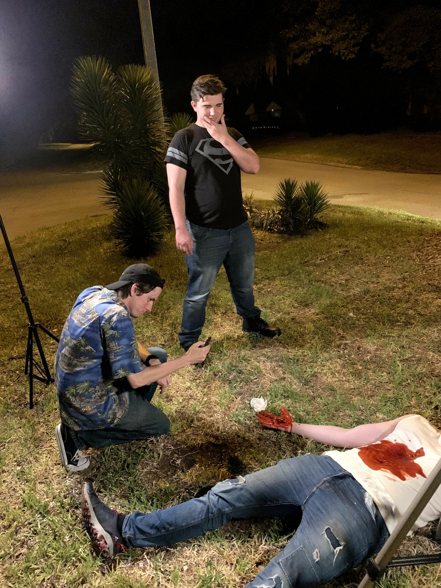 Two young men outdoors at night, one sitting on the ground with a phone, the other standing near a chair with a blood-stained shirt, grass, and bushes in background.