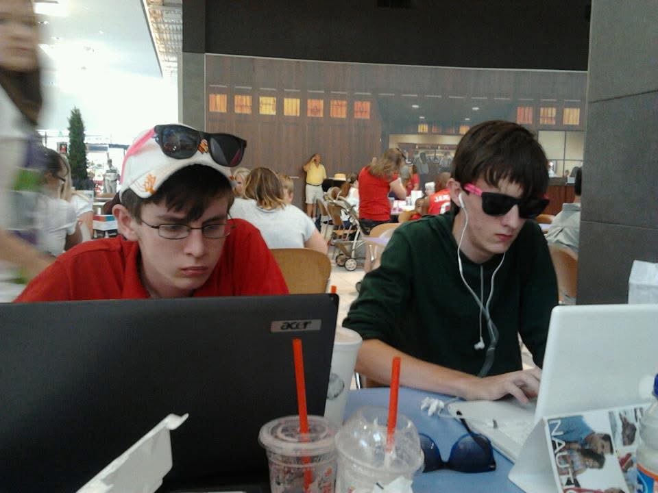 Two young men sit at a table with laptops, wearing sunglasses, in a busy indoor setting.