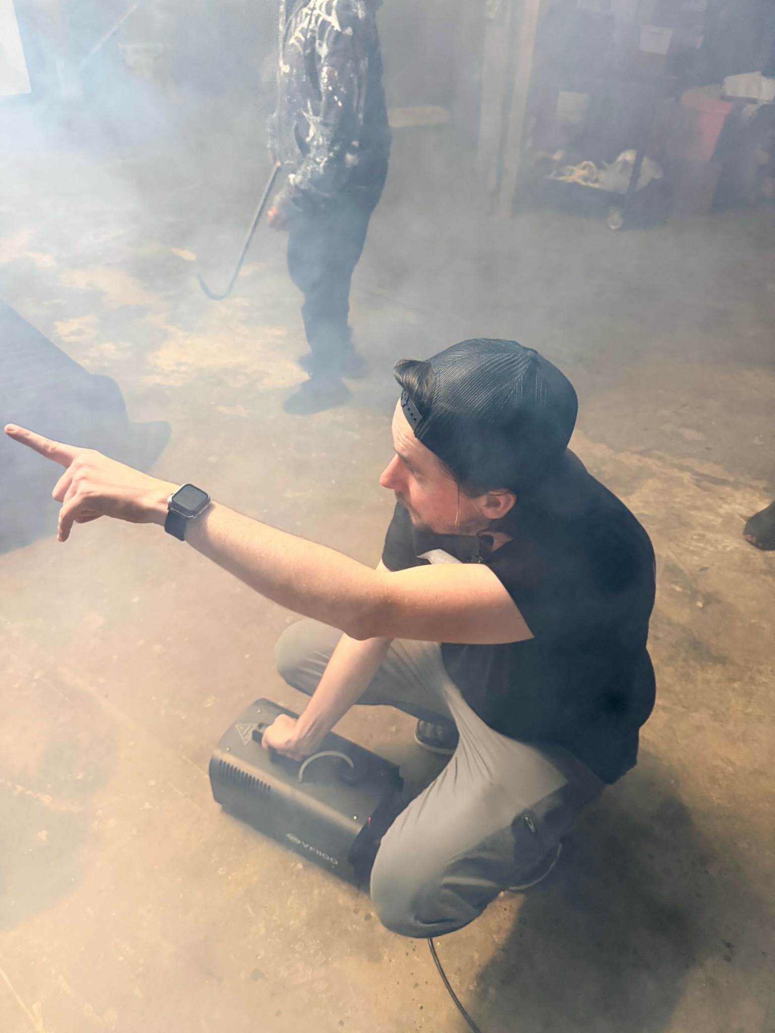 Person crouching on the ground, pointing with their right arm, surrounded by smoke or fog, wearing a black cap and shirt.