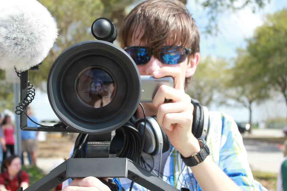Person with sunglasses holding a camera outdoors, with trees and sky in background.