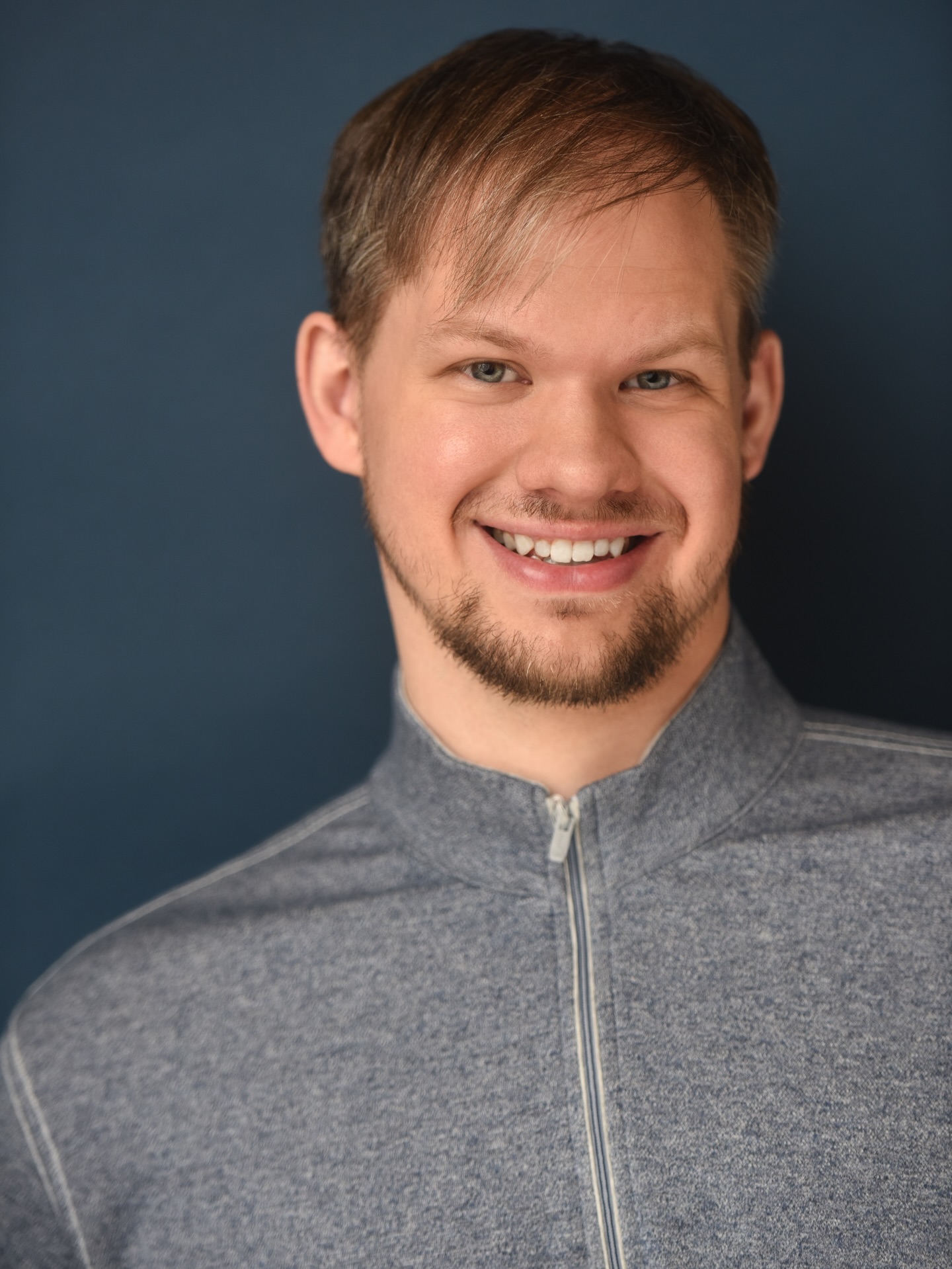 Smiling man with short light brown hair and a beard, wearing a gray zip-up jacket, against a dark background.