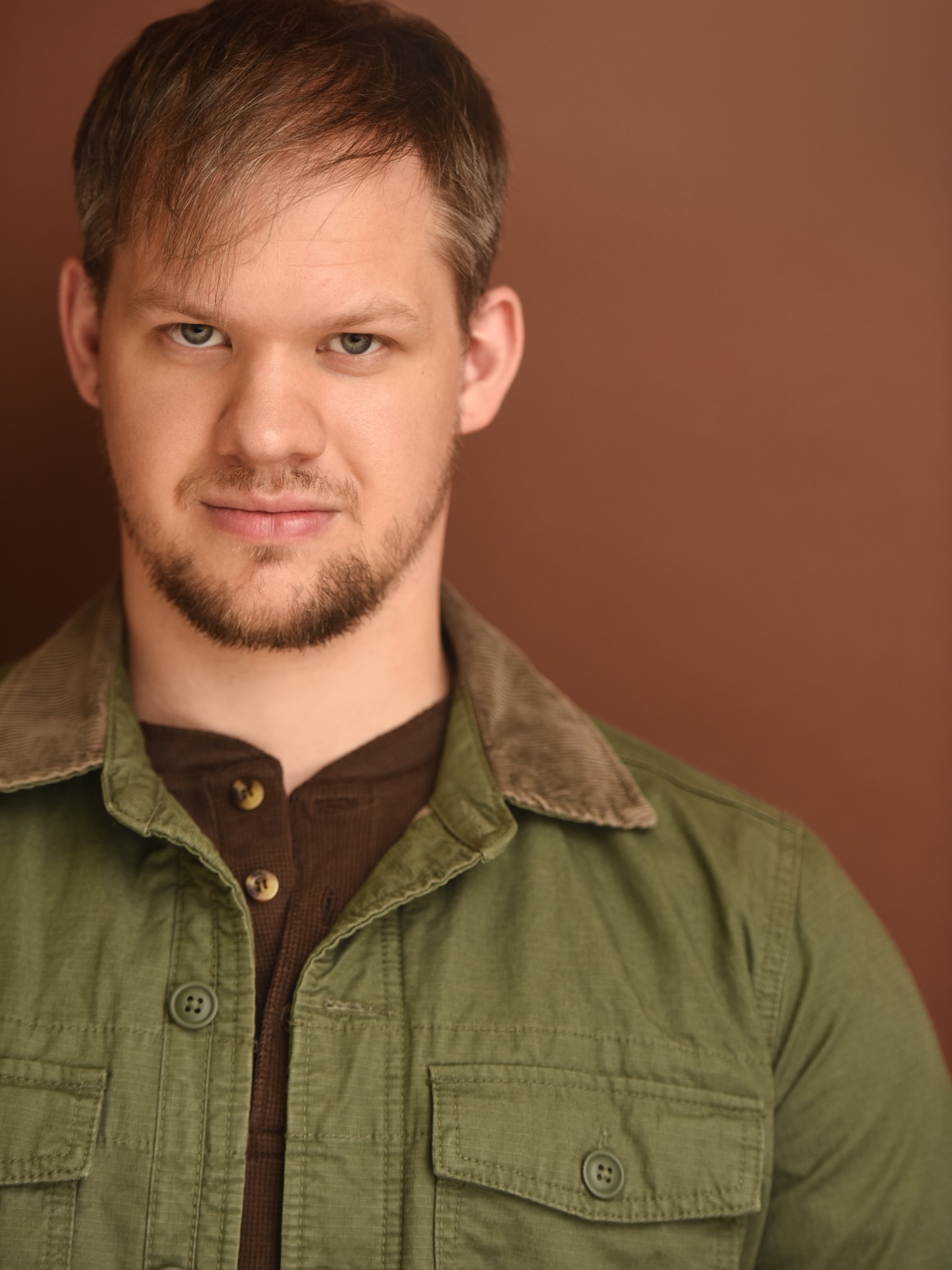 Portrait of a young man with short brown hair and a beard, wearing a green jacket, against a brown background.