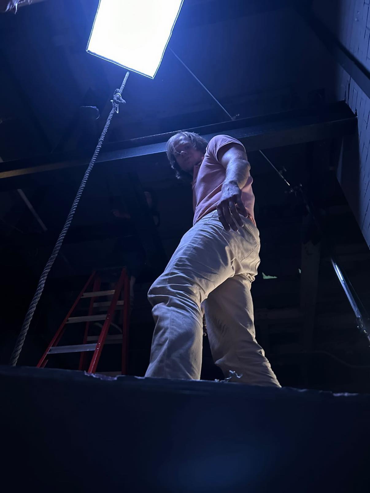 Person standing on a dark stage with a bright light above, looking down at the camera, low angle view.