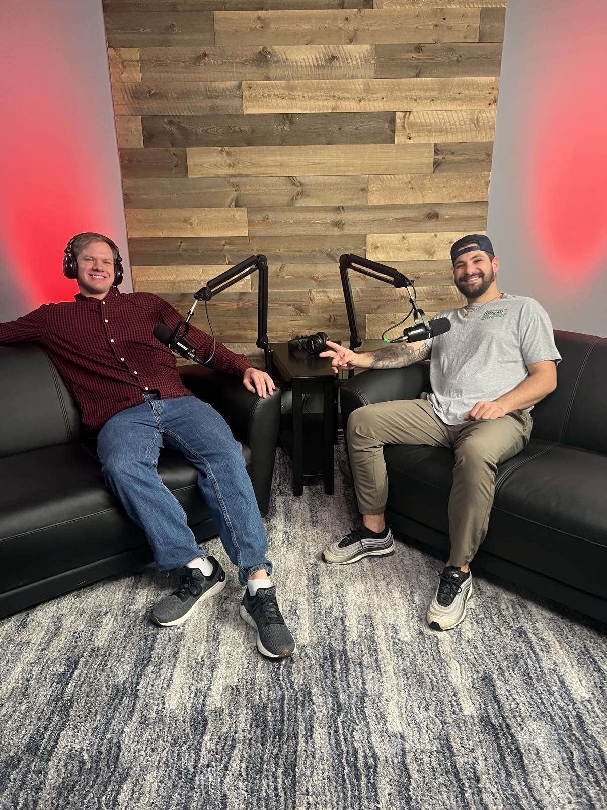 Two men sit on black couches in a recording studio with wooden wall panels and red lighting, shaking hands.