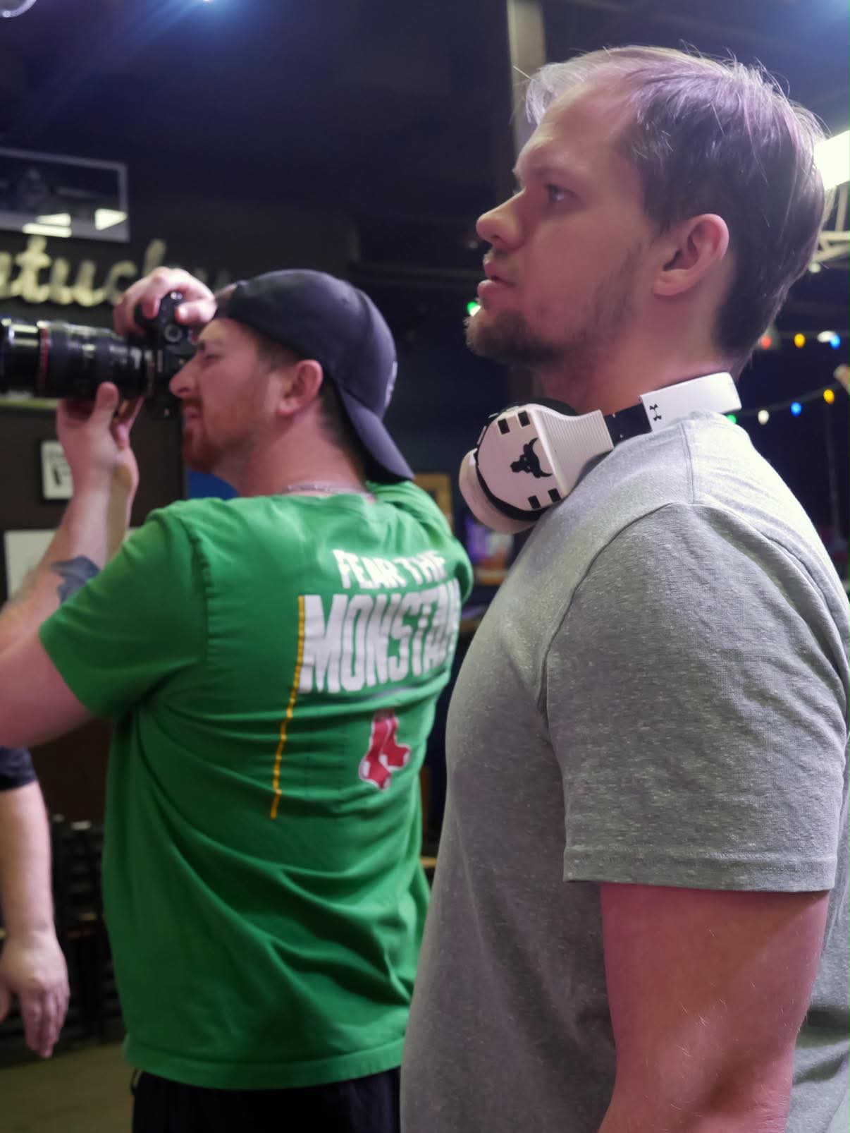 Two men in a bar, one drinking from a bottle, the other wearing headphones around his neck, both facing right.