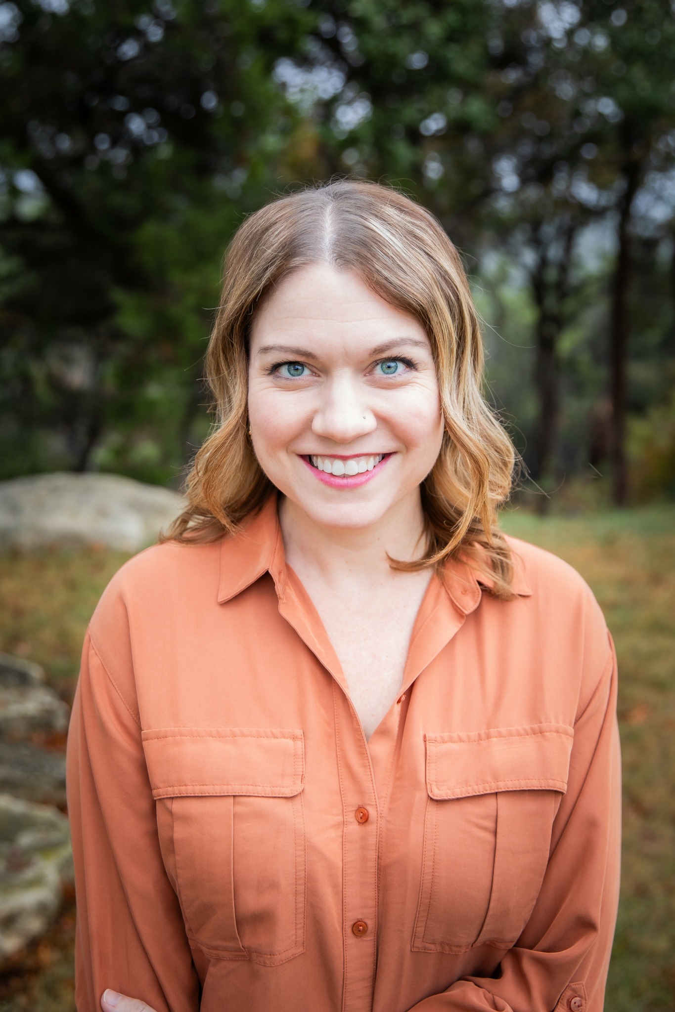 Young woman with shoulder-length wavy hair smiling outdoors, wearing an orange shirt, trees and rocks in background.