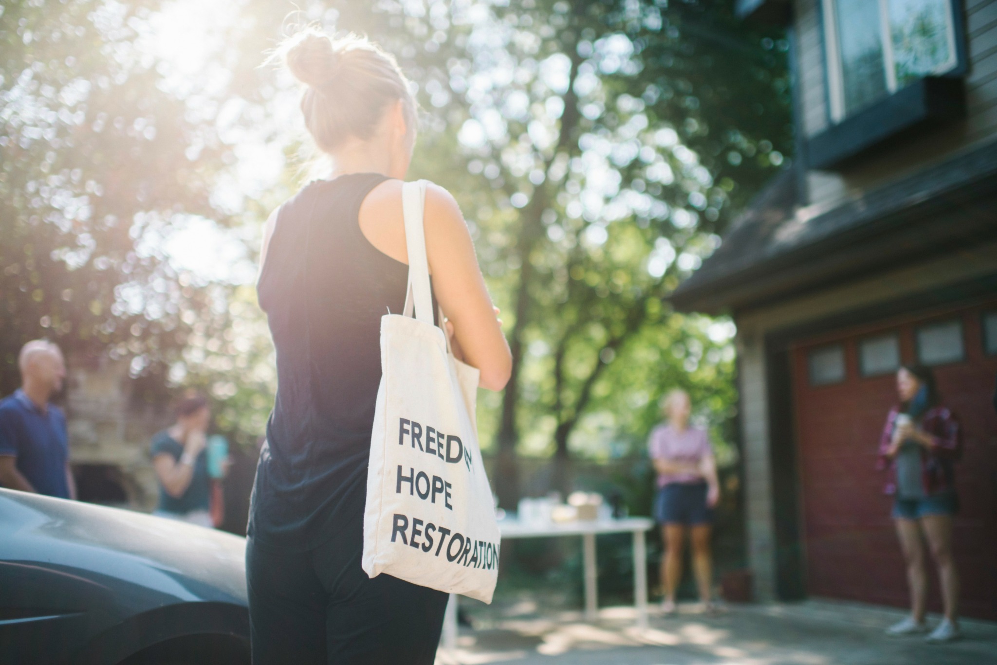 Back view of a woman with a tote bag, outdoors with trees and people in the background, sunlight shining through.
