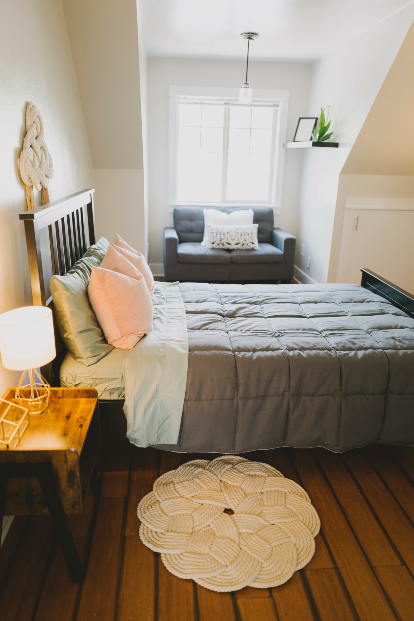 Bedroom with bed, pillows, nightstand, window, and a small sofa under the window, with a round rug on wooden floor.