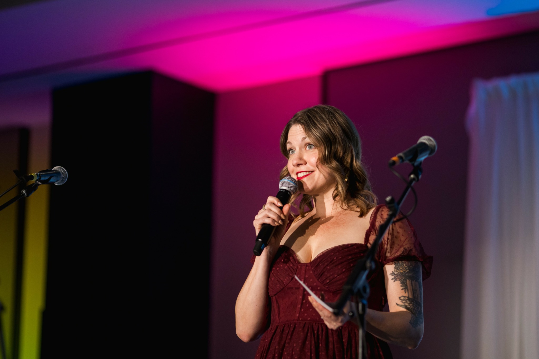 Woman with shoulder-length hair speaking into a microphone on stage, holding a paper, with colorful lighting in background.