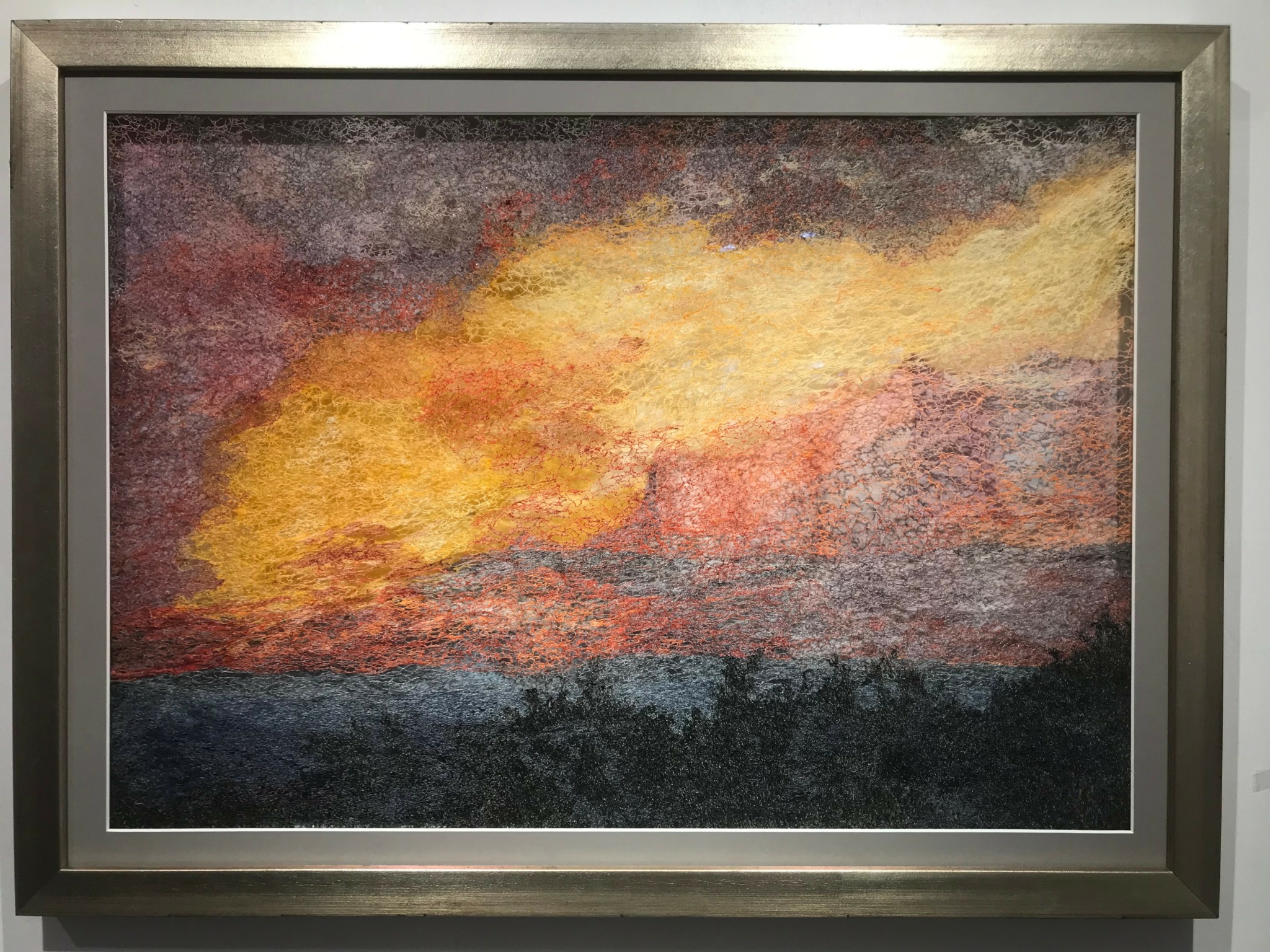 Painting of a colorful sunset sky with clouds over a dark landscape, in a silver frame.