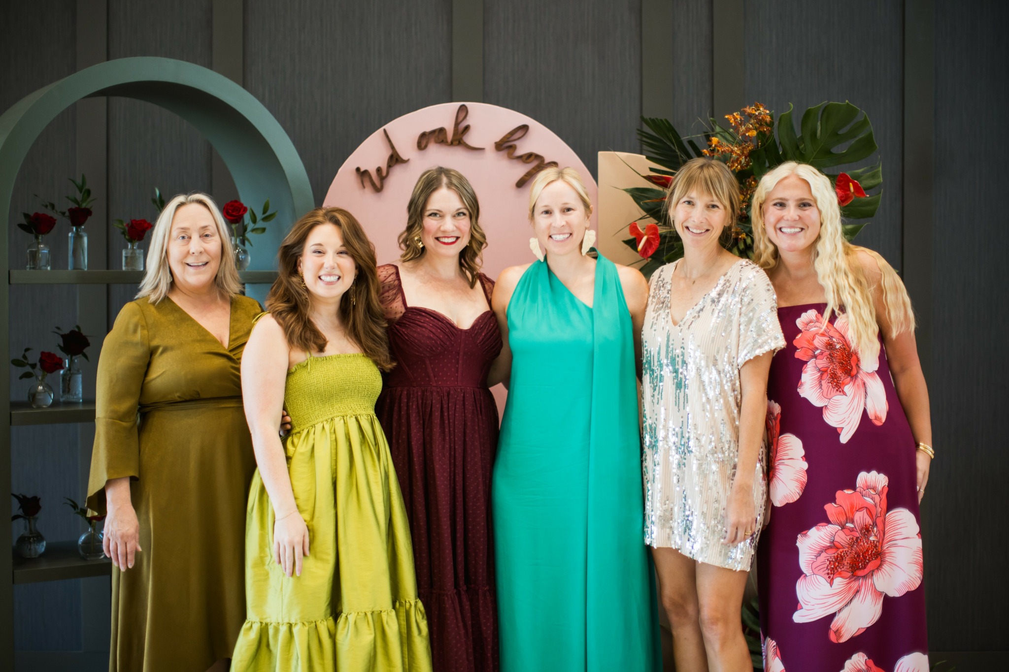Six women standing together indoors, smiling, with a decorative background and flowers, celebrating a special occasion.