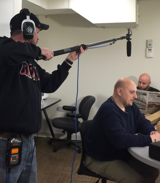 Person holding a boom microphone over a seated man reading a newspaper in an office, with another person in the background. 20 words.