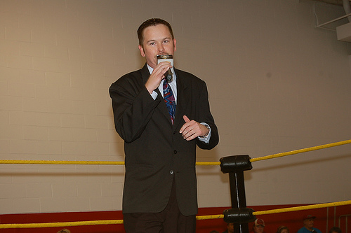 Man in a suit speaking into a microphone at an indoor event, with a yellow railing behind him.