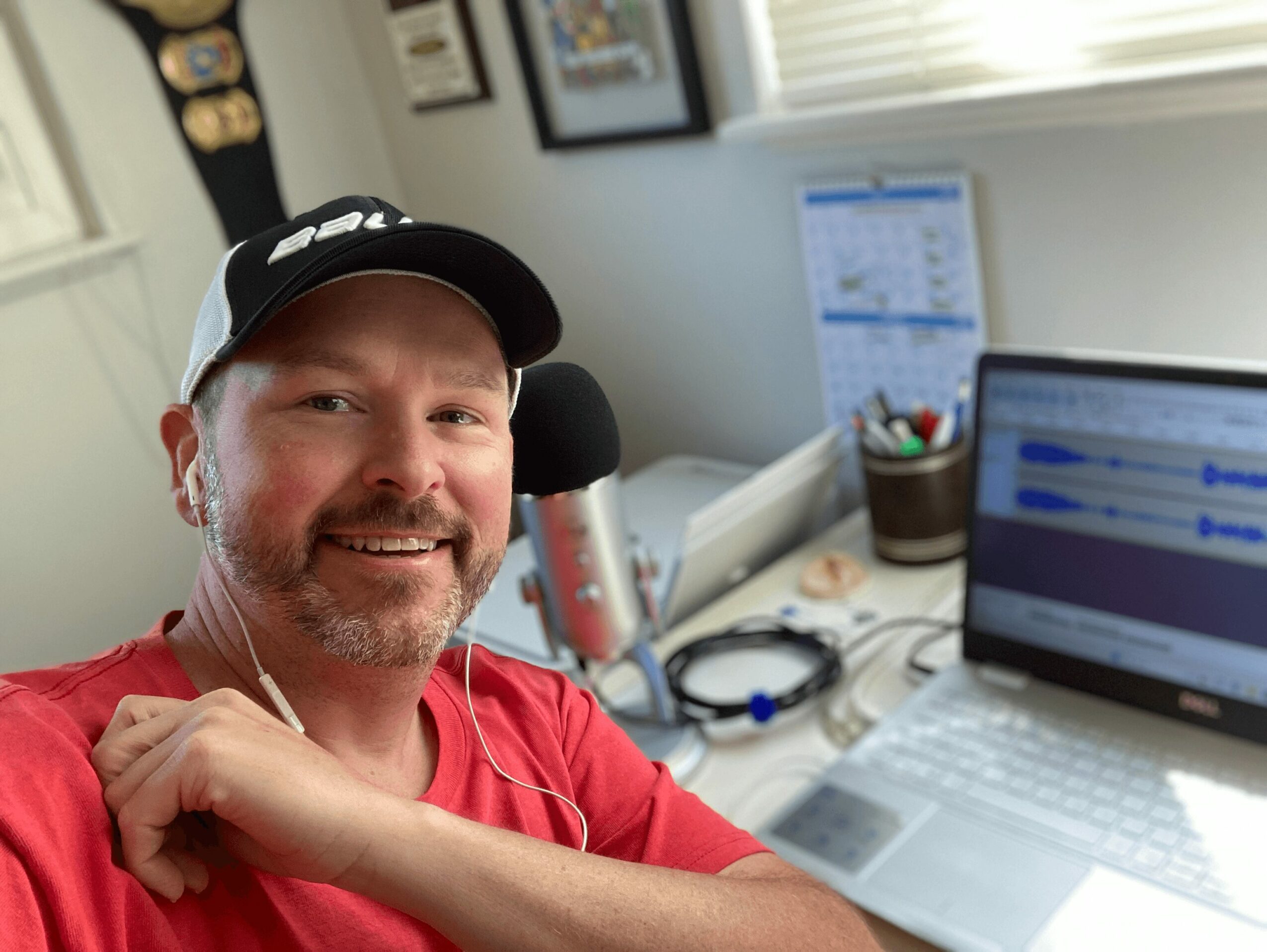Man with a beard and cap smiling at a desk with a laptop, microphone, and office supplies.