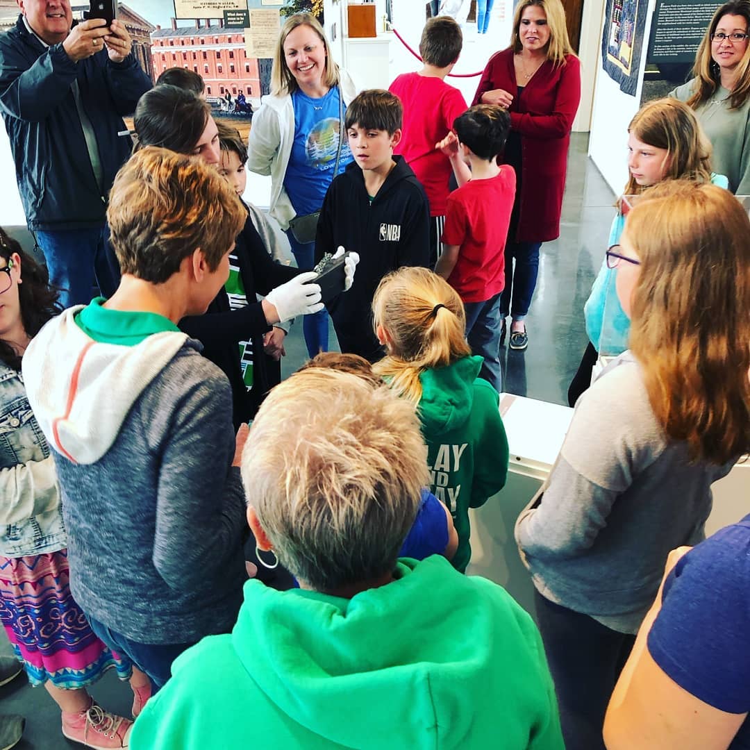 Group of children and adults gathered indoors, some children wearing colorful clothing, engaging in conversation or activity, with a cityscape visible through windows.