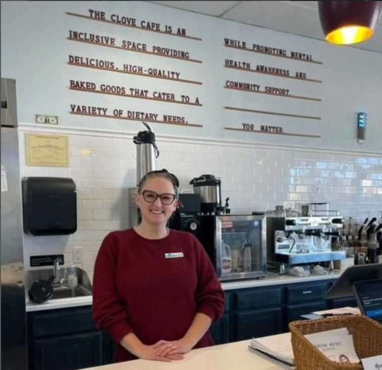 Smiling woman in red sweater behind counter in cafe, white tiled wall with text in background, basket on counter.