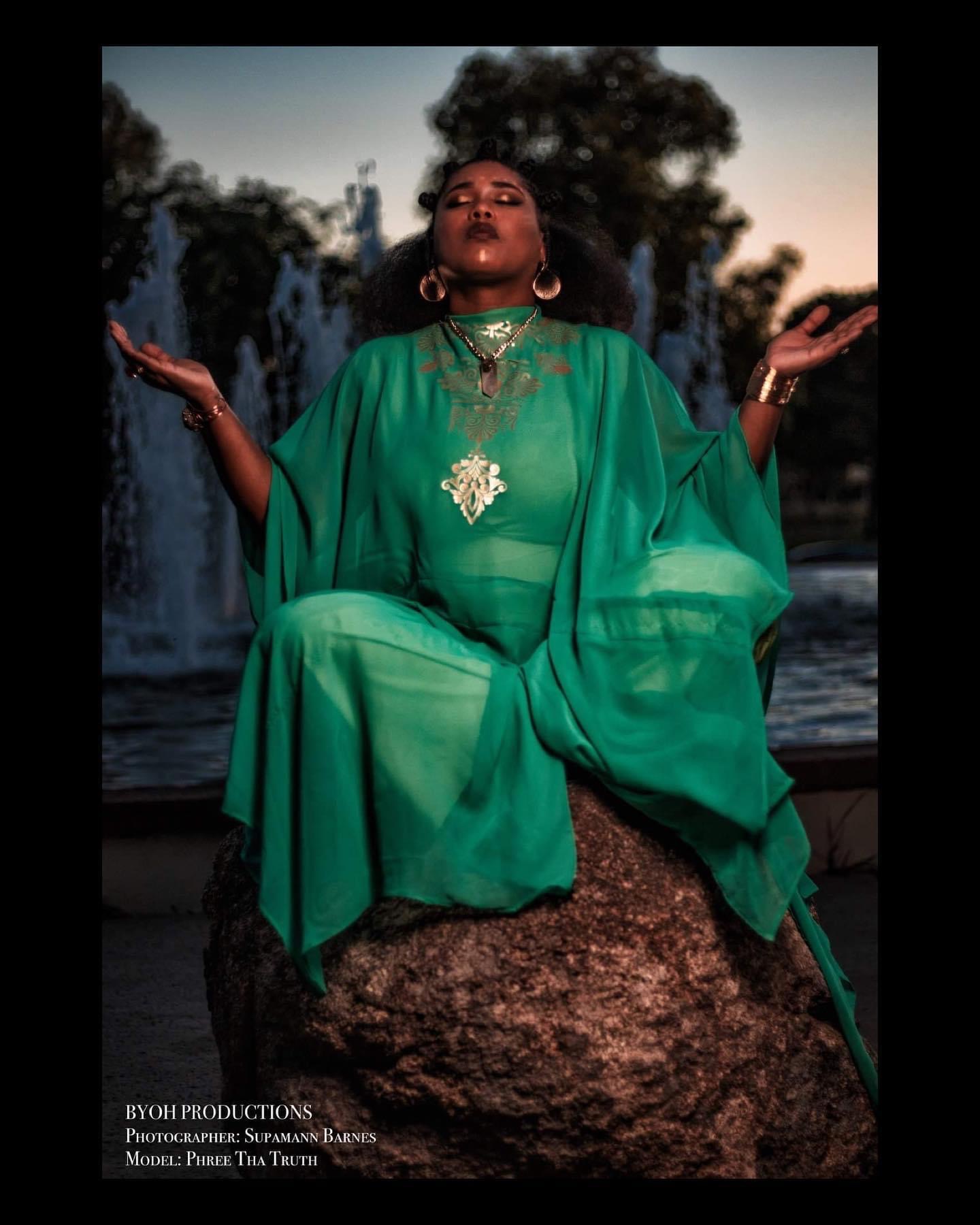 Woman in green dress with jewelry, sitting on a rock near water fountain, with trees and sky in background.